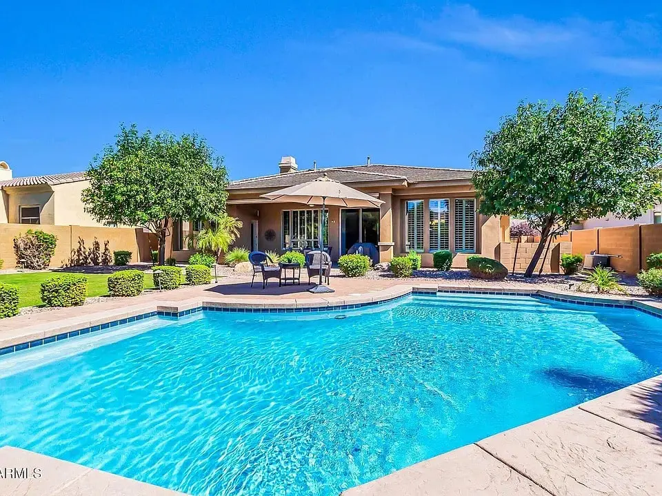 Backyard with a swimming pool, patio furniture, and trees under a clear blue sky.