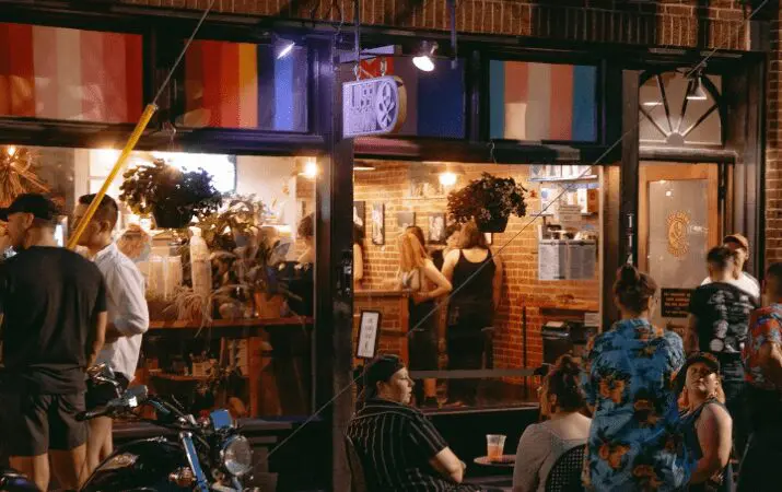 People socializing outside a bar with rainbow flags displayed above the entrance.