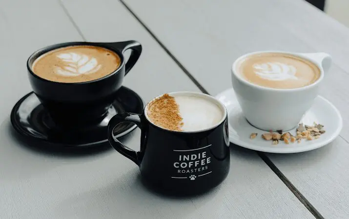 Three cups of latte art coffee on a table, one cup labeled 'Indie Coffee Roasters'.