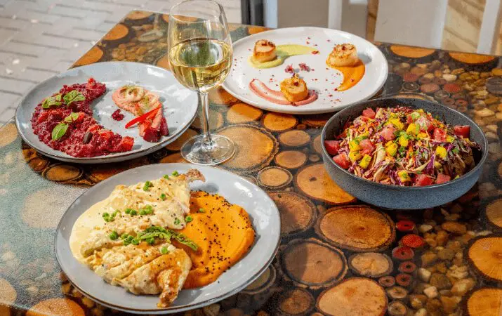 A table with four gourmet dishes and a glass of white wine on a wooden surface.