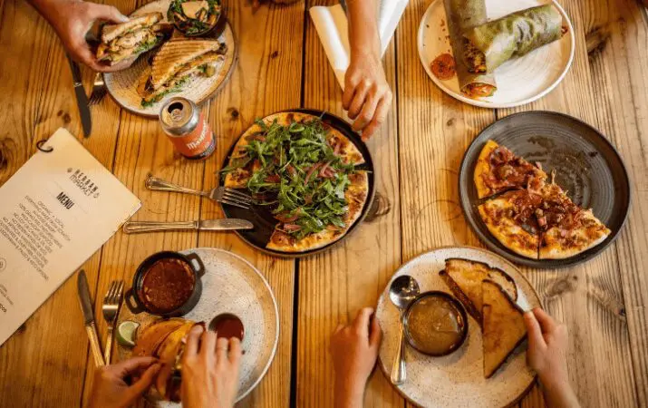 Aerial view of people sharing various pizzas and sandwiches at a wooden table.