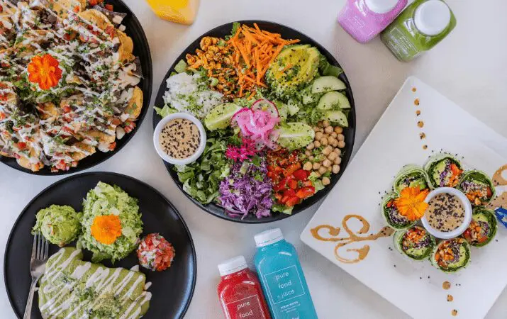 A variety of colorful vegan dishes and juices arranged on a table, featuring salads and sushi.