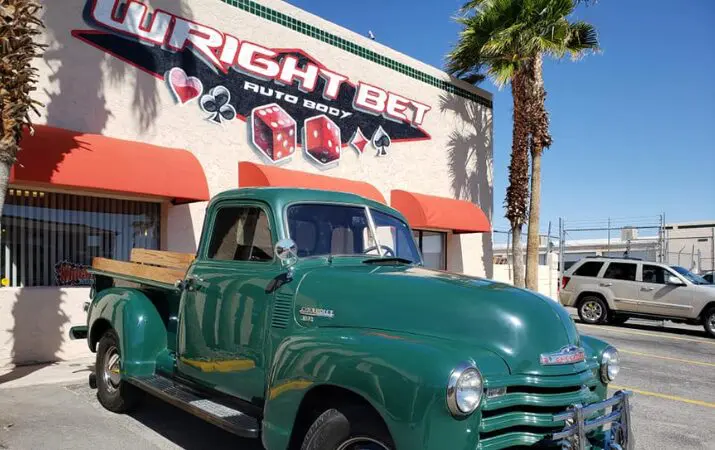 Green vintage truck parked in front of 'Wright Bet Auto Body' shop with palm trees nearby.