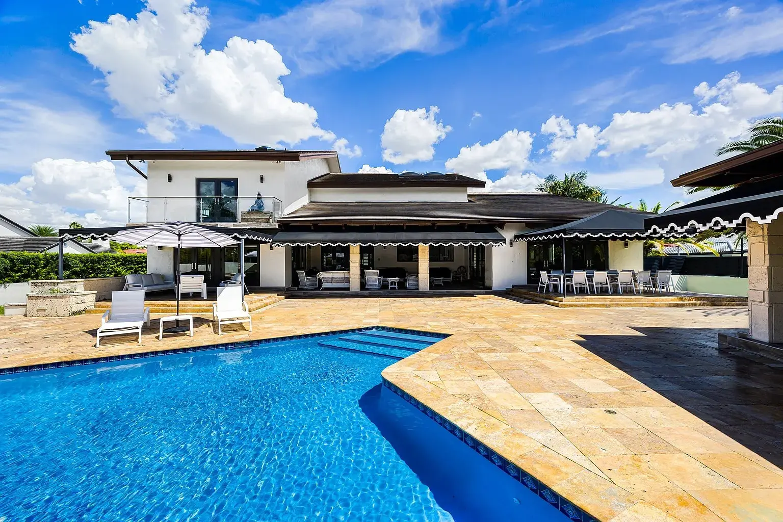 Luxurious modern house with a large patio, swimming pool, and outdoor seating under a blue sky.