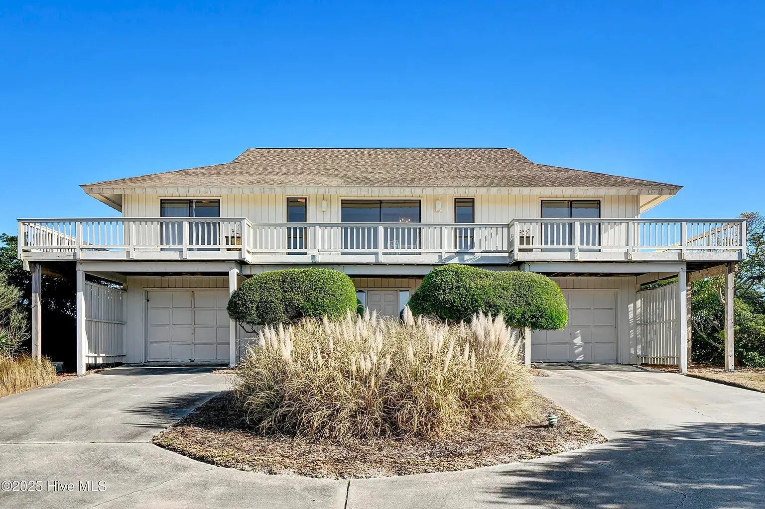 Two-story house with wraparound balcony, manicured shrubs, and a double garage under a clear blue sky.
