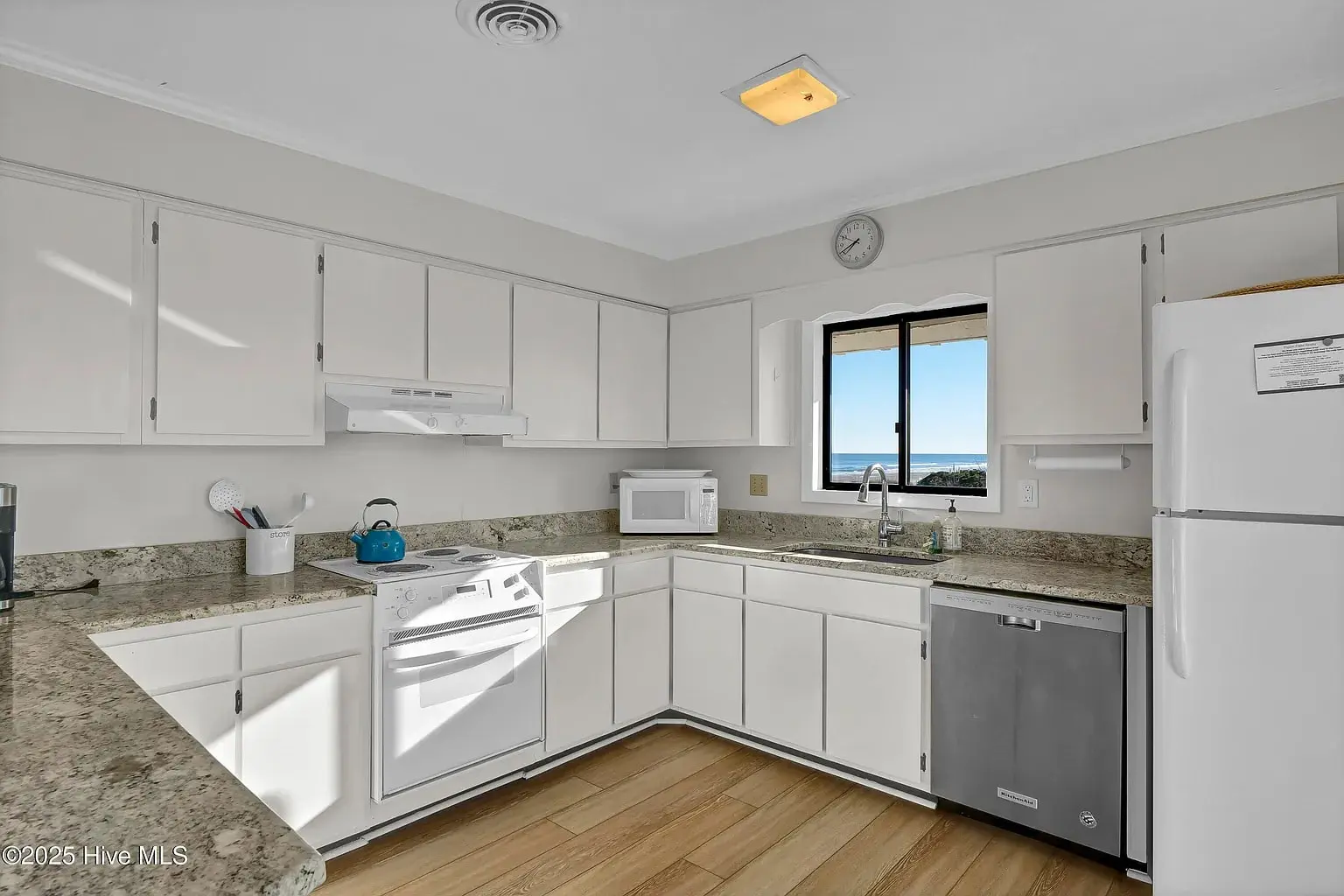 Modern kitchen with white cabinets, granite countertops, and ocean view through window.
