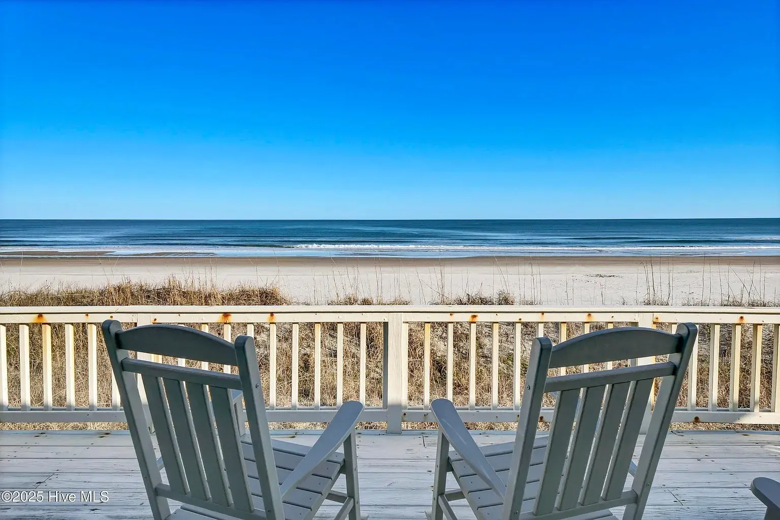 Two rocking chairs on a deck overlooking a sandy beach and calm ocean waves.