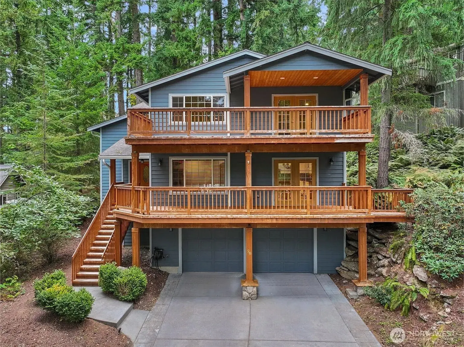 Two-story blue house with wooden balconies, surrounded by tall trees and greenery.