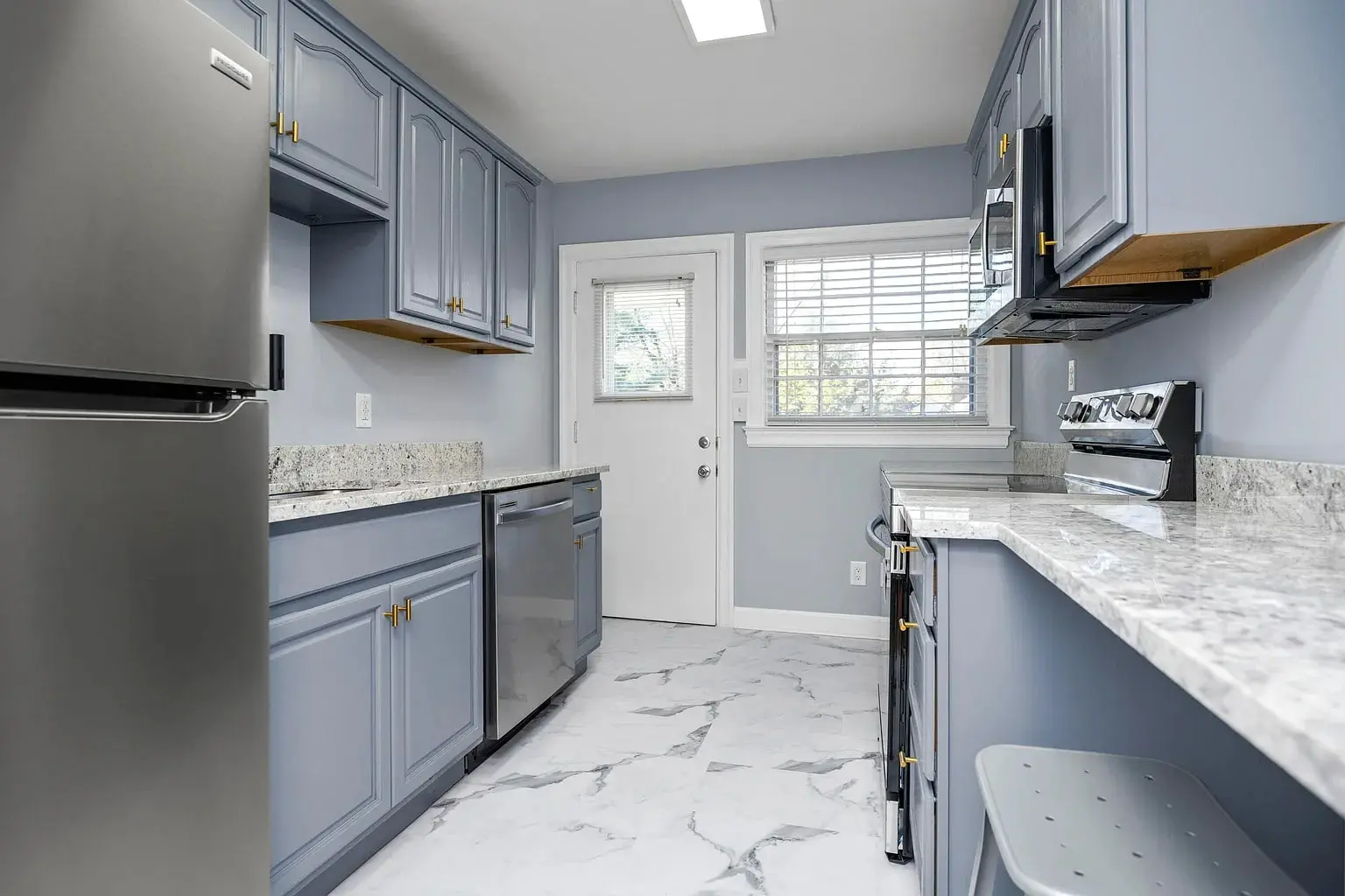 Modern kitchen with gray cabinets, marble countertops, stainless steel appliances, and white tile flooring.