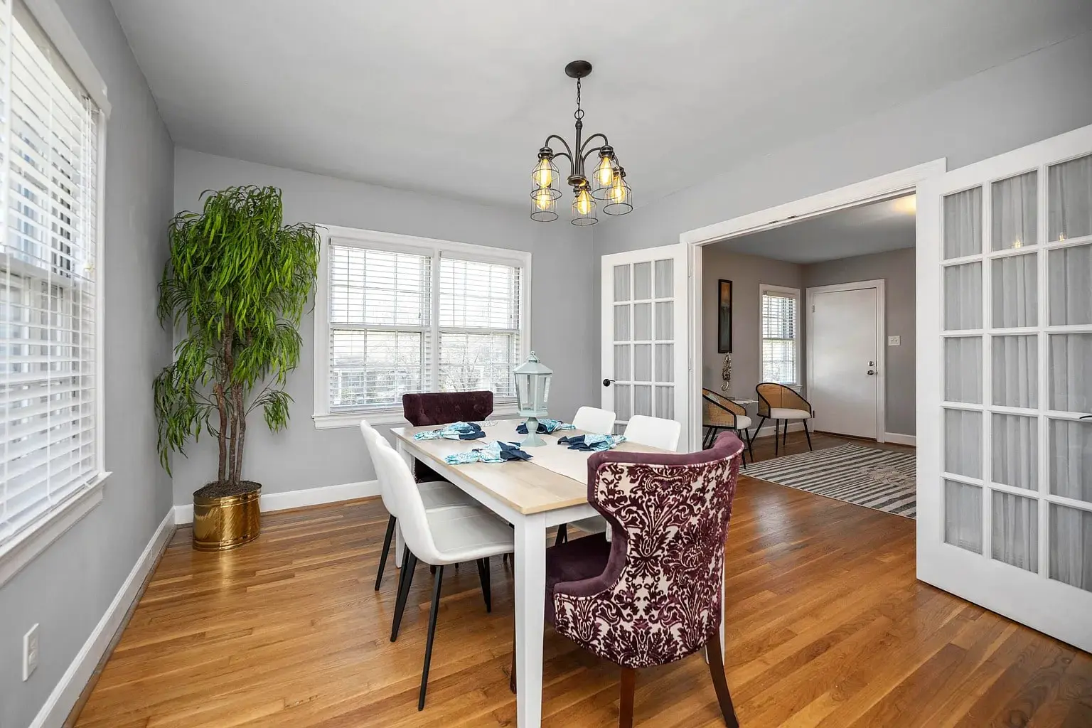 Dining room with a wooden table, white and patterned chairs, chandelier, and large potted plant.
