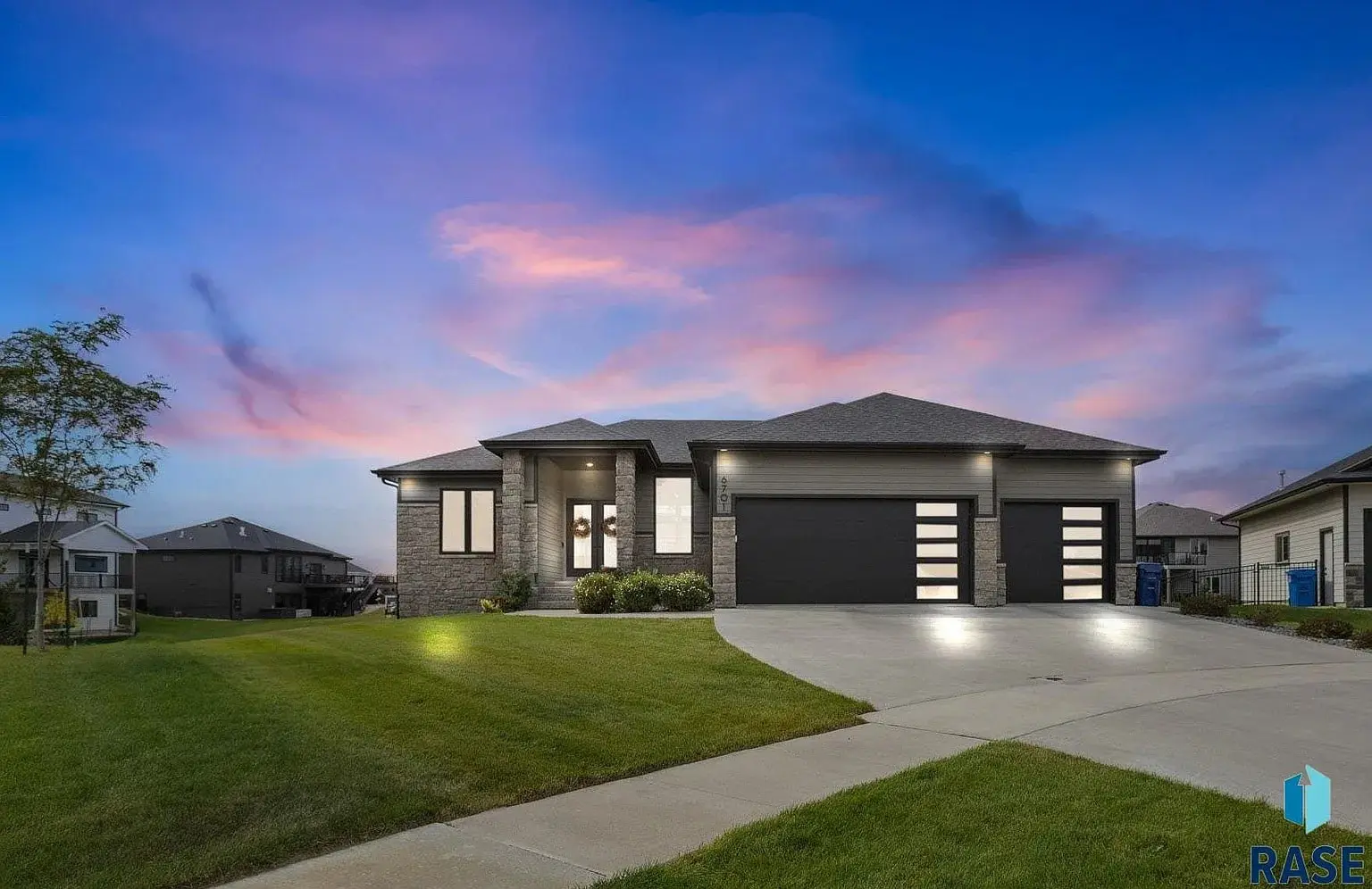 Modern single-story house with a large driveway, lit exterior, and a vibrant sunset sky.