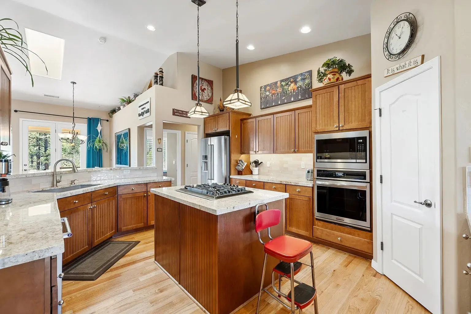 Modern kitchen with wooden cabinets, central island, red stool, stainless steel appliances, and decorative accents.