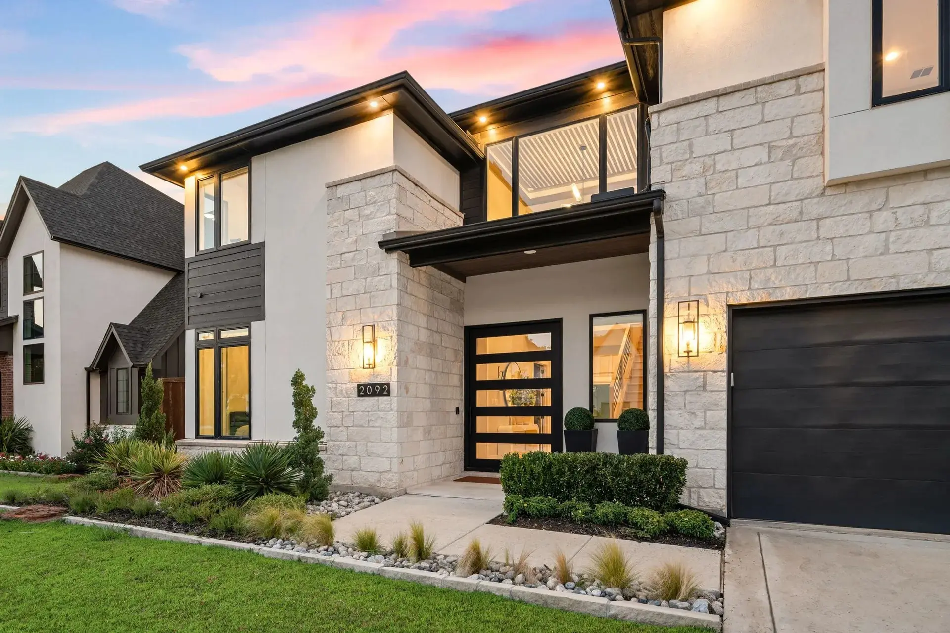Modern two-story house with stone facade, large windows, and a well-maintained lawn at sunset.