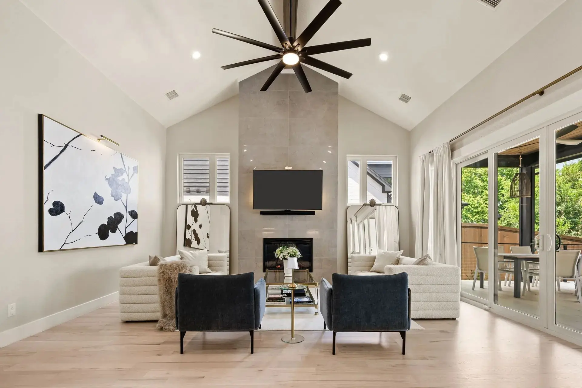 Modern living room with high ceiling, large fan, abstract art, and patio view through glass doors.