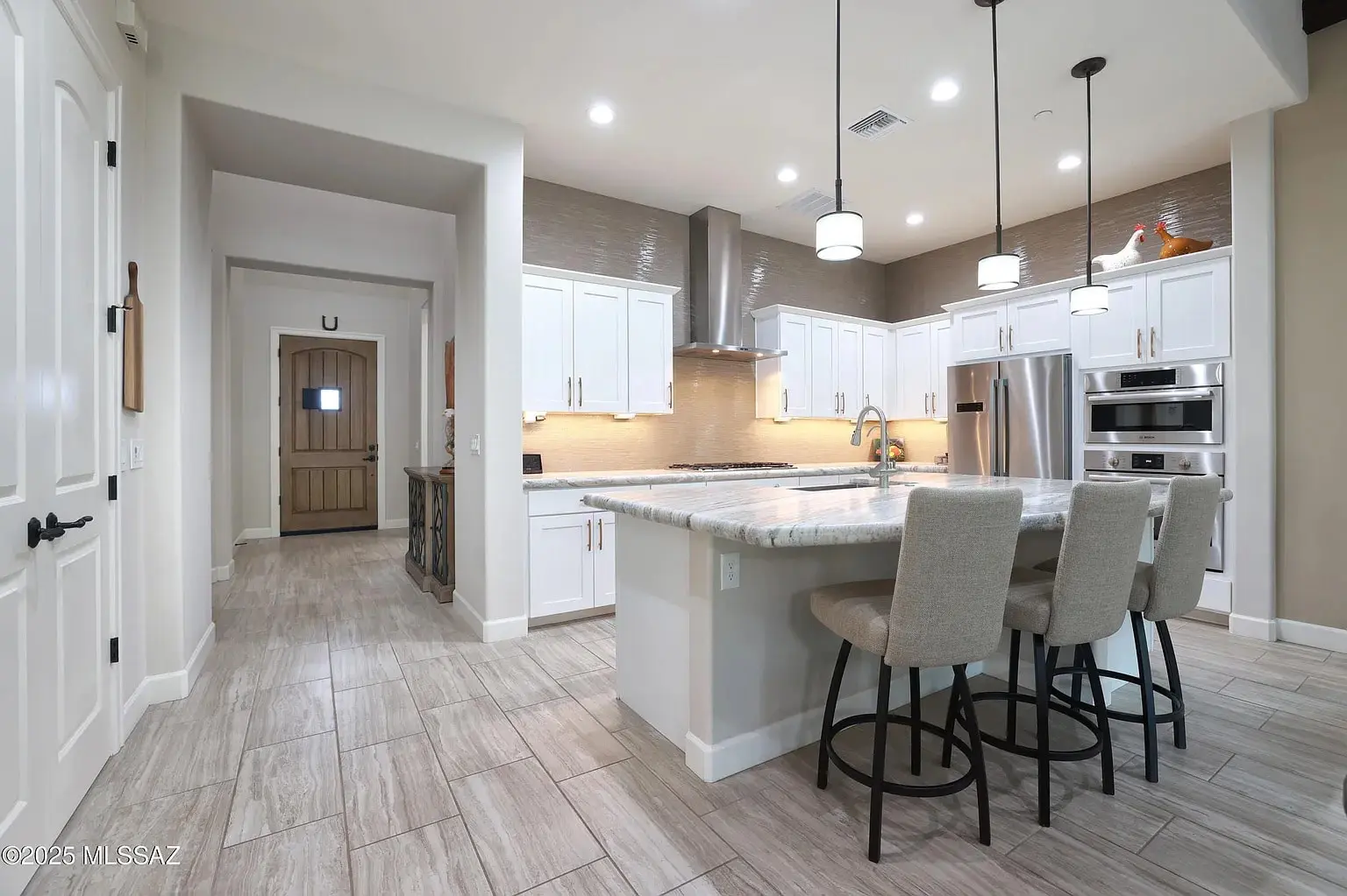 Modern kitchen with white cabinets, marble island, bar stools, stainless steel appliances, and wood-look tile flooring.