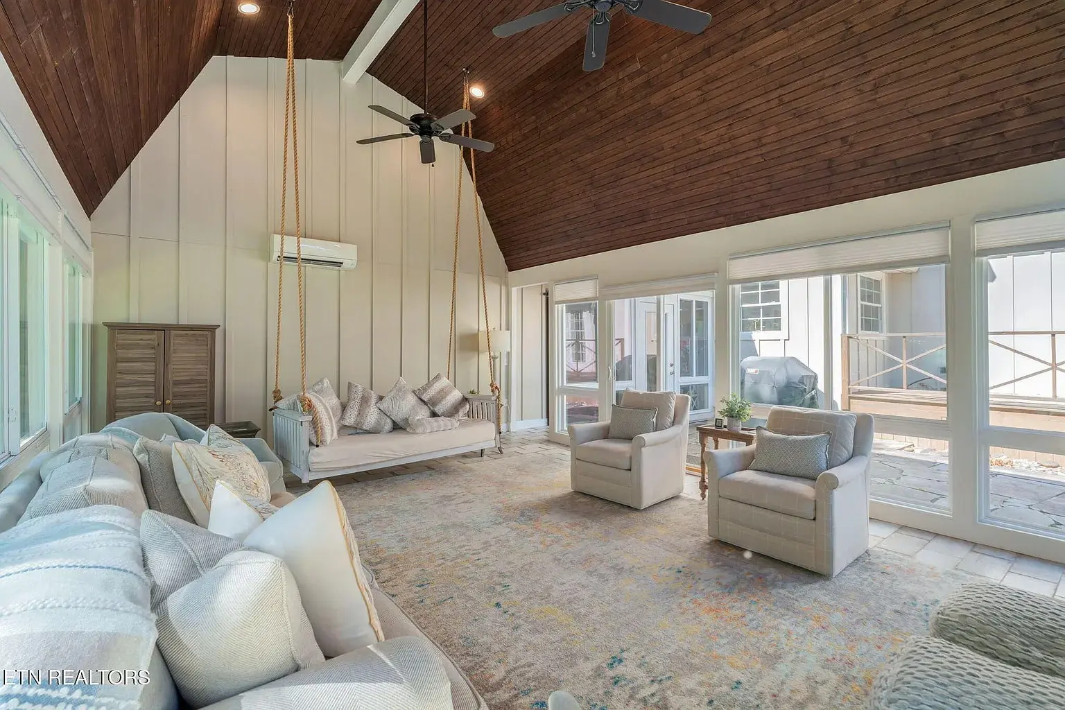 Spacious living room with vaulted wooden ceiling, large windows, sofas, armchairs, and a hanging daybed.