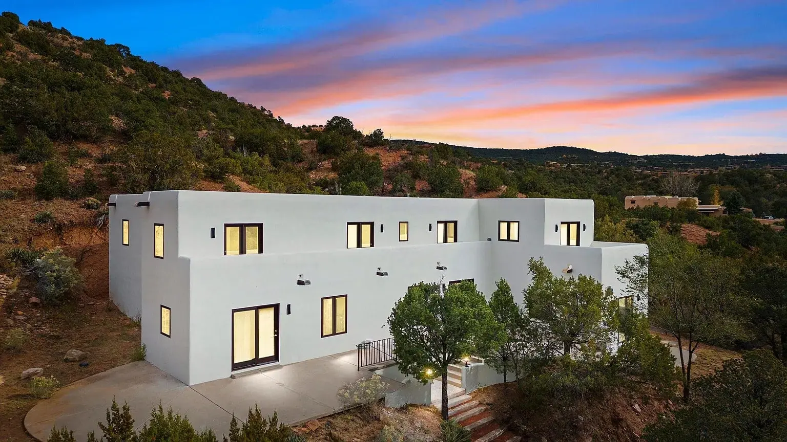 Modern white house with large windows, surrounded by trees, set against a colorful sunset sky.