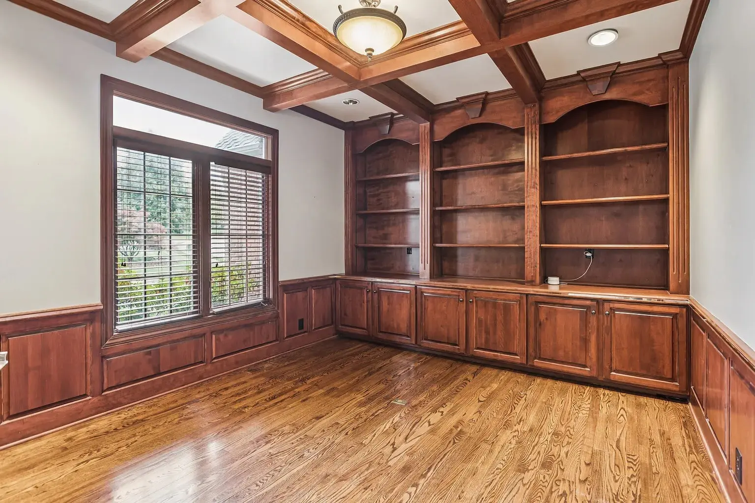 Empty room with wooden floors, built-in wooden shelves, and large window with blinds.