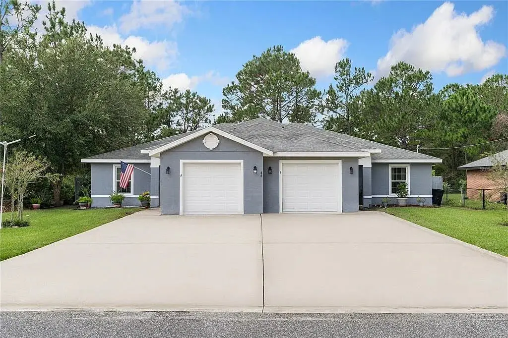 Gray duplex with two garages, American flag, surrounded by trees and grass, under a blue sky.