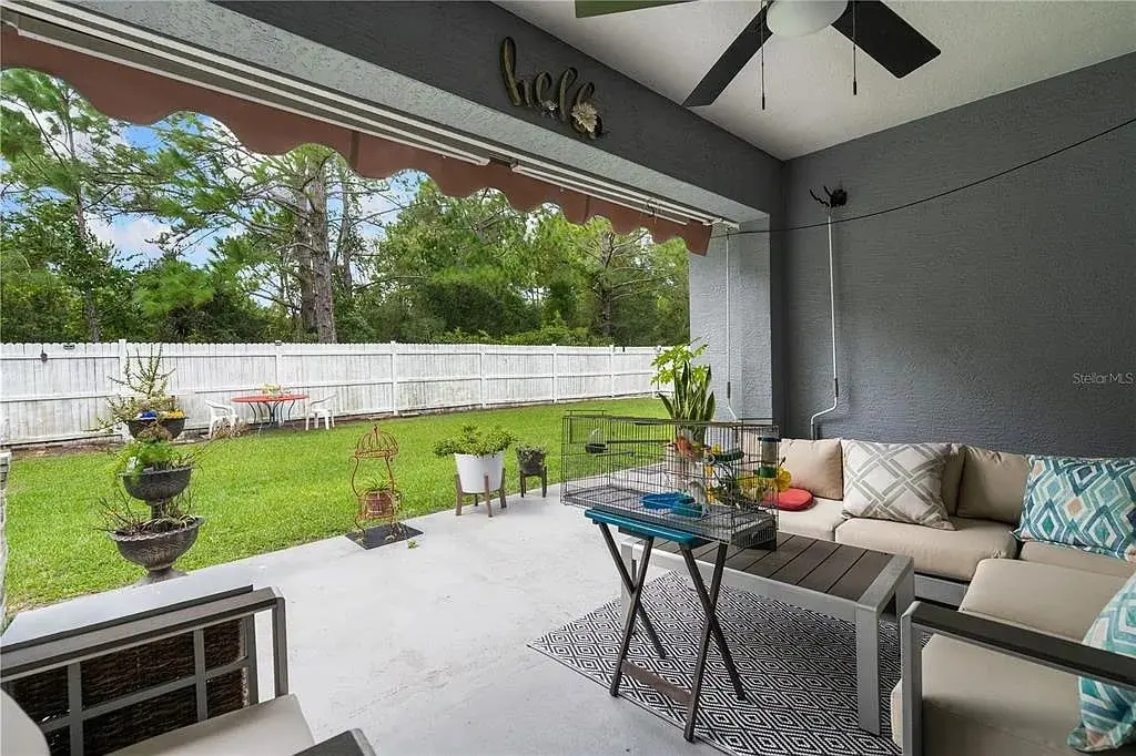 Covered patio with seating, table, and plants, overlooking a grassy yard with trees and a white fence.