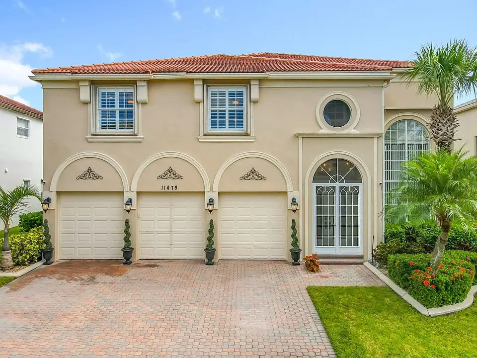 Two-story beige house with three-car garage, red-tiled roof, and landscaped front yard.