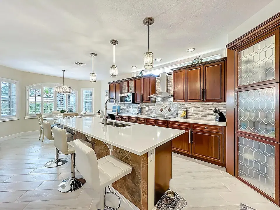 Modern kitchen with wooden cabinets, white countertops, island seating, pendant lights, and large windows.
