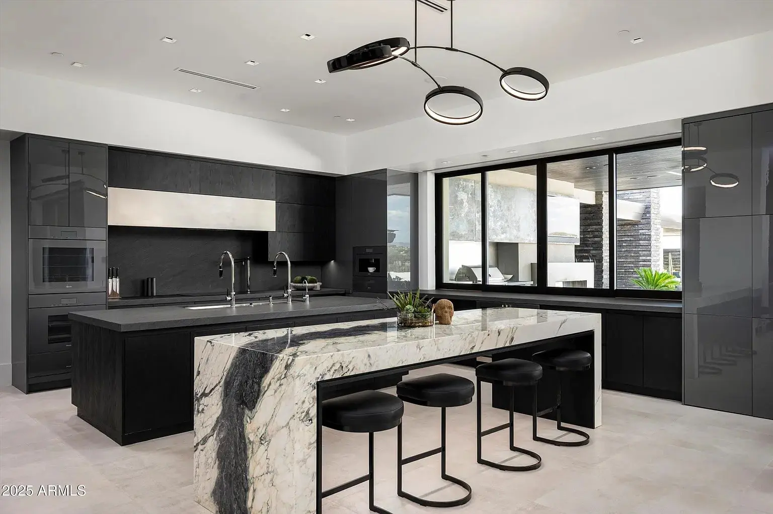 Modern kitchen with black cabinetry, marble island, and contemporary lighting fixtures. Large windows provide natural light.