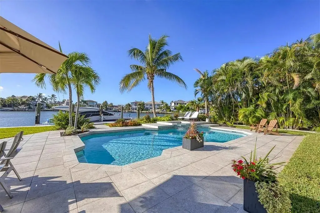 Outdoor pool area with palm trees, overlooking a waterfront with boats and clear blue sky.