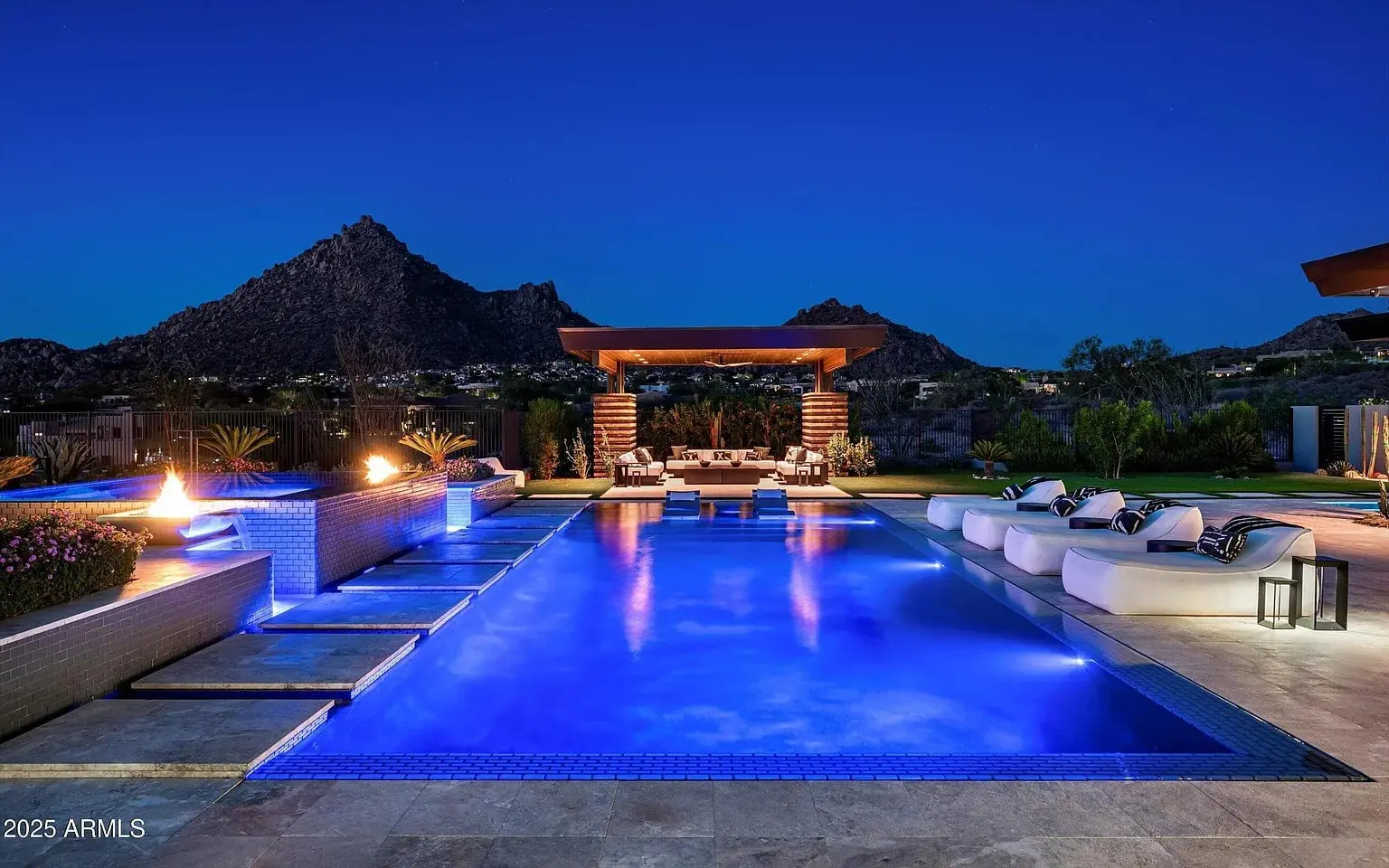 Luxurious outdoor pool area at night with fire pit, lounge chairs, and mountain view.