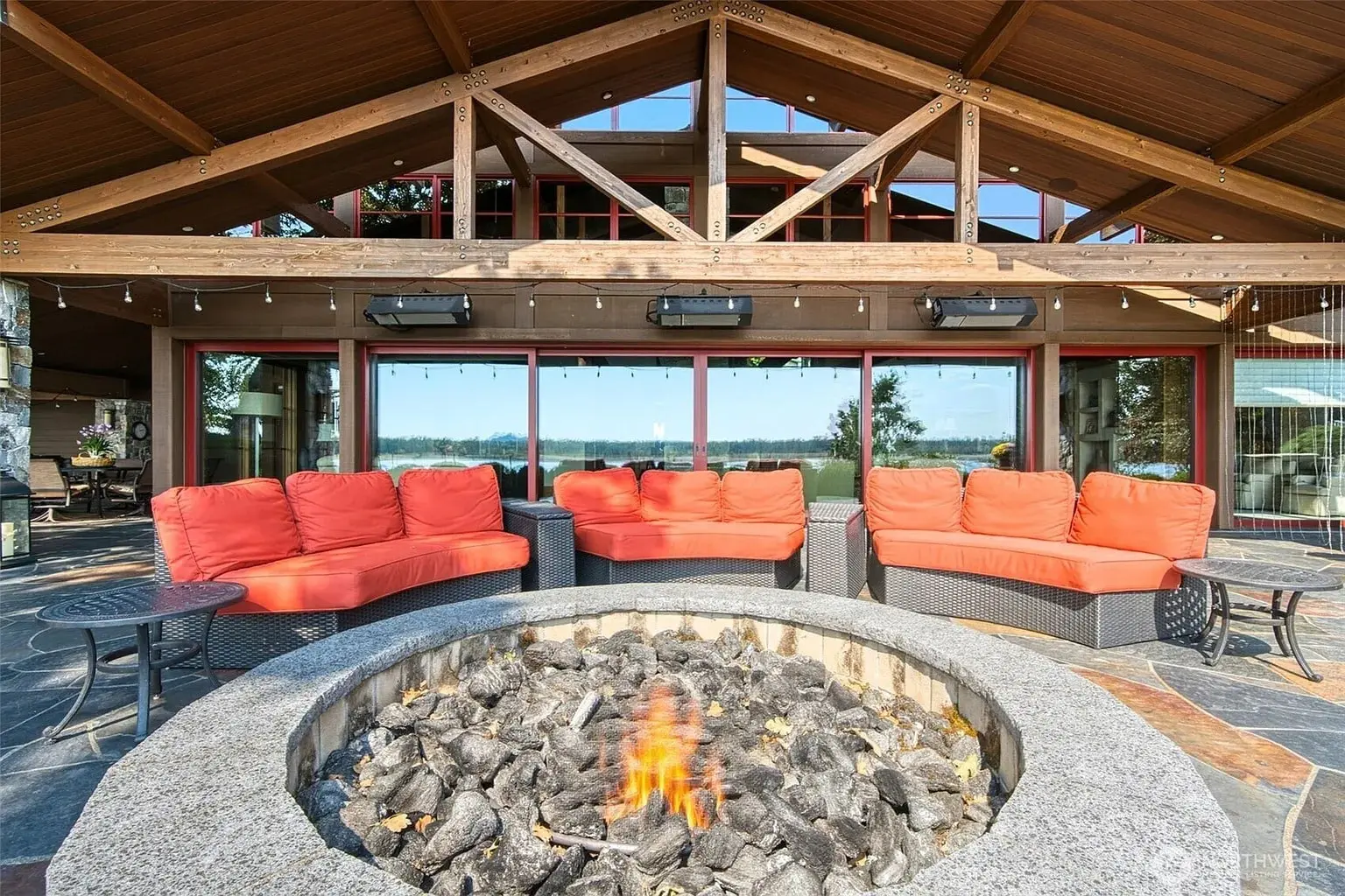 Outdoor patio with red-cushioned seating around a stone fire pit, under a wooden roof.