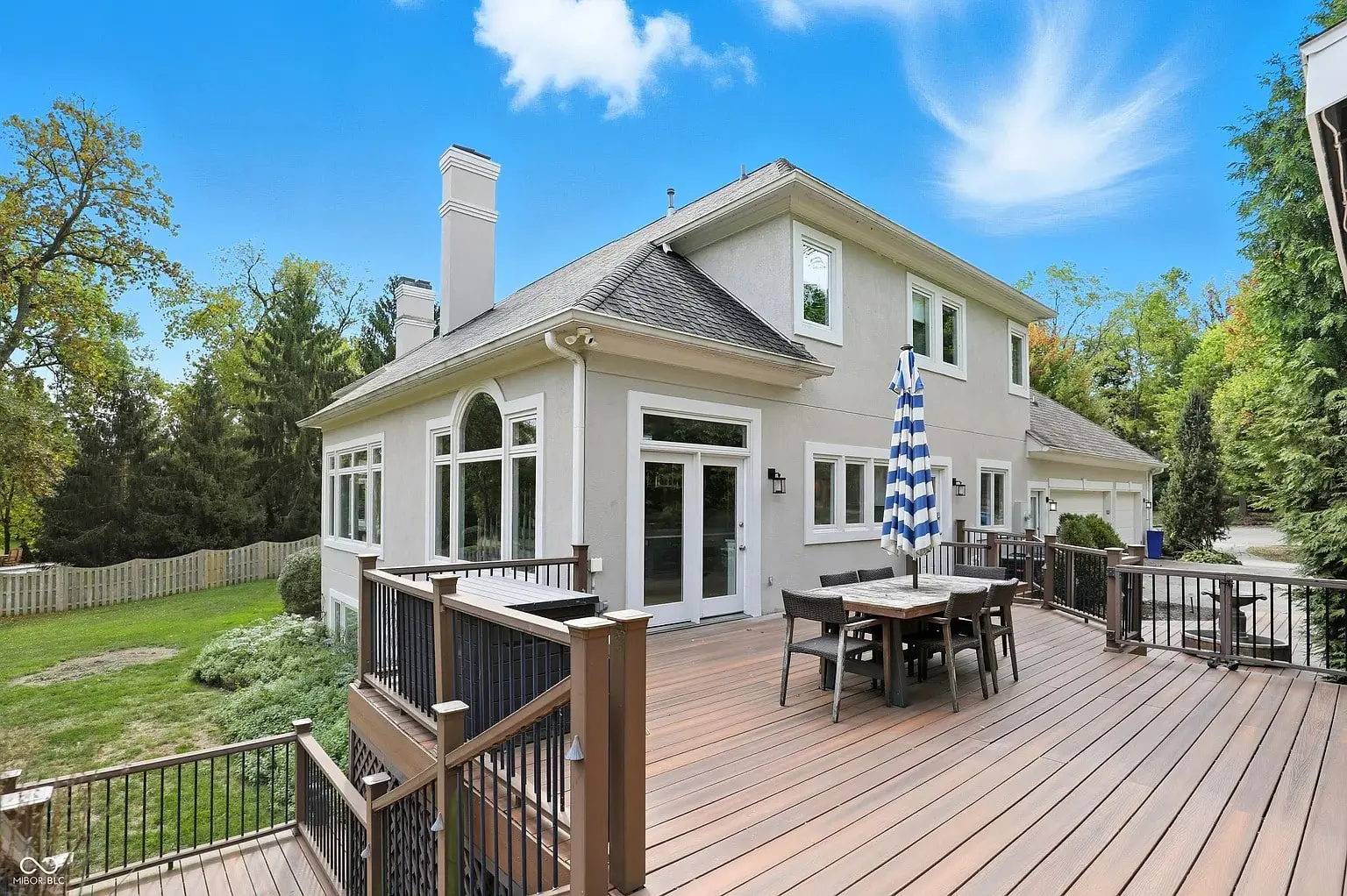 Two-story house with large wooden deck, outdoor dining set, and umbrella, surrounded by trees.