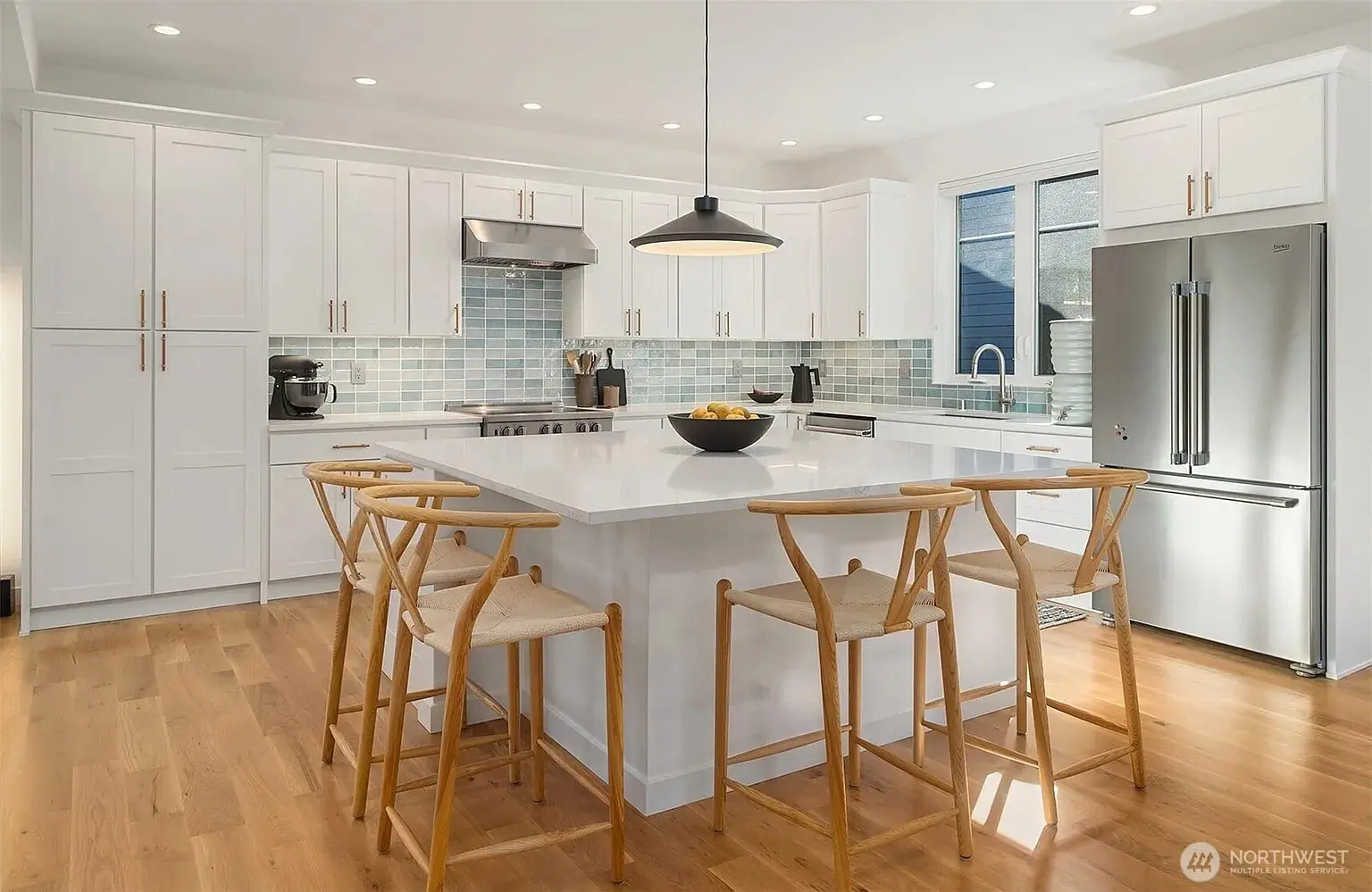 Modern kitchen with white cabinets, island, wooden chairs, stainless steel appliances, and light wood flooring.