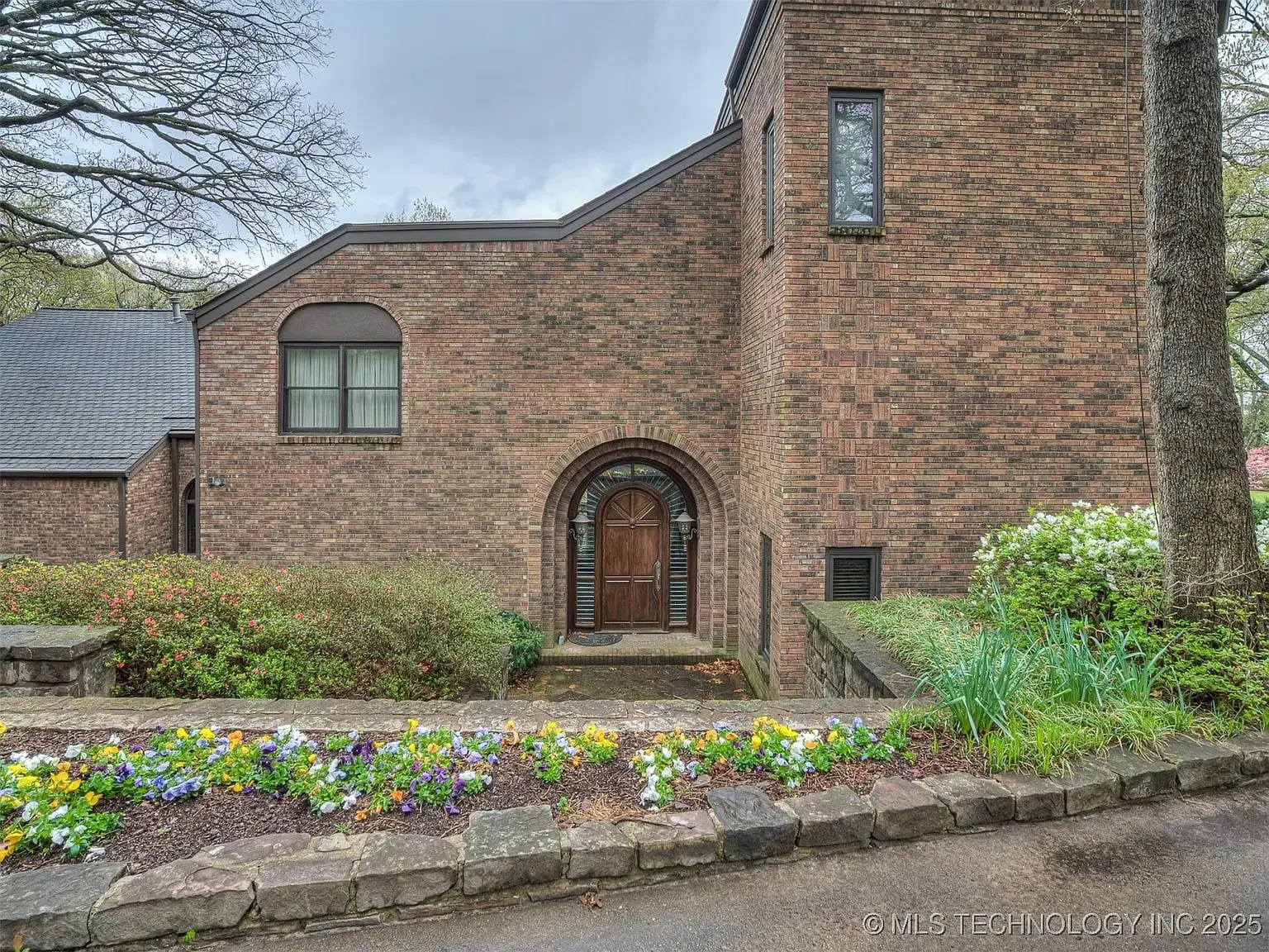 Brick house with arched wooden door, surrounded by colorful flowers and greenery.