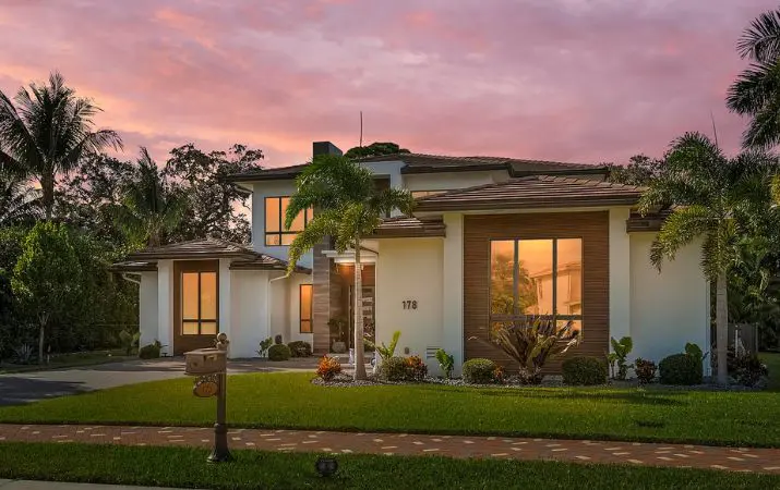Modern two-story house with large windows, surrounded by palm trees, at sunset.