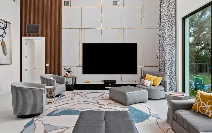 Modern living room with large TV, abstract rug, gray seating, and floor-to-ceiling windows.