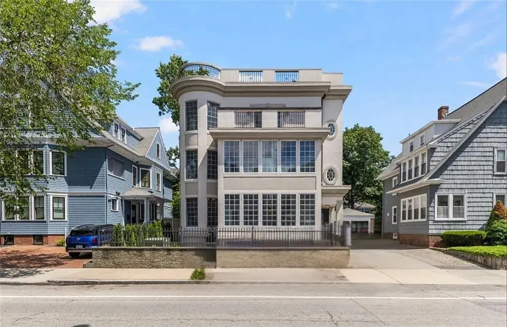 Modern three-story house with large windows, flanked by traditional homes on a suburban street.