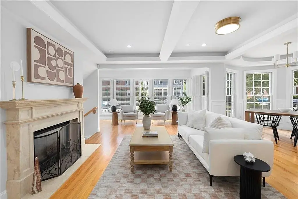 Spacious living room with white sofa, fireplace, abstract art, and large windows. Dining area visible.