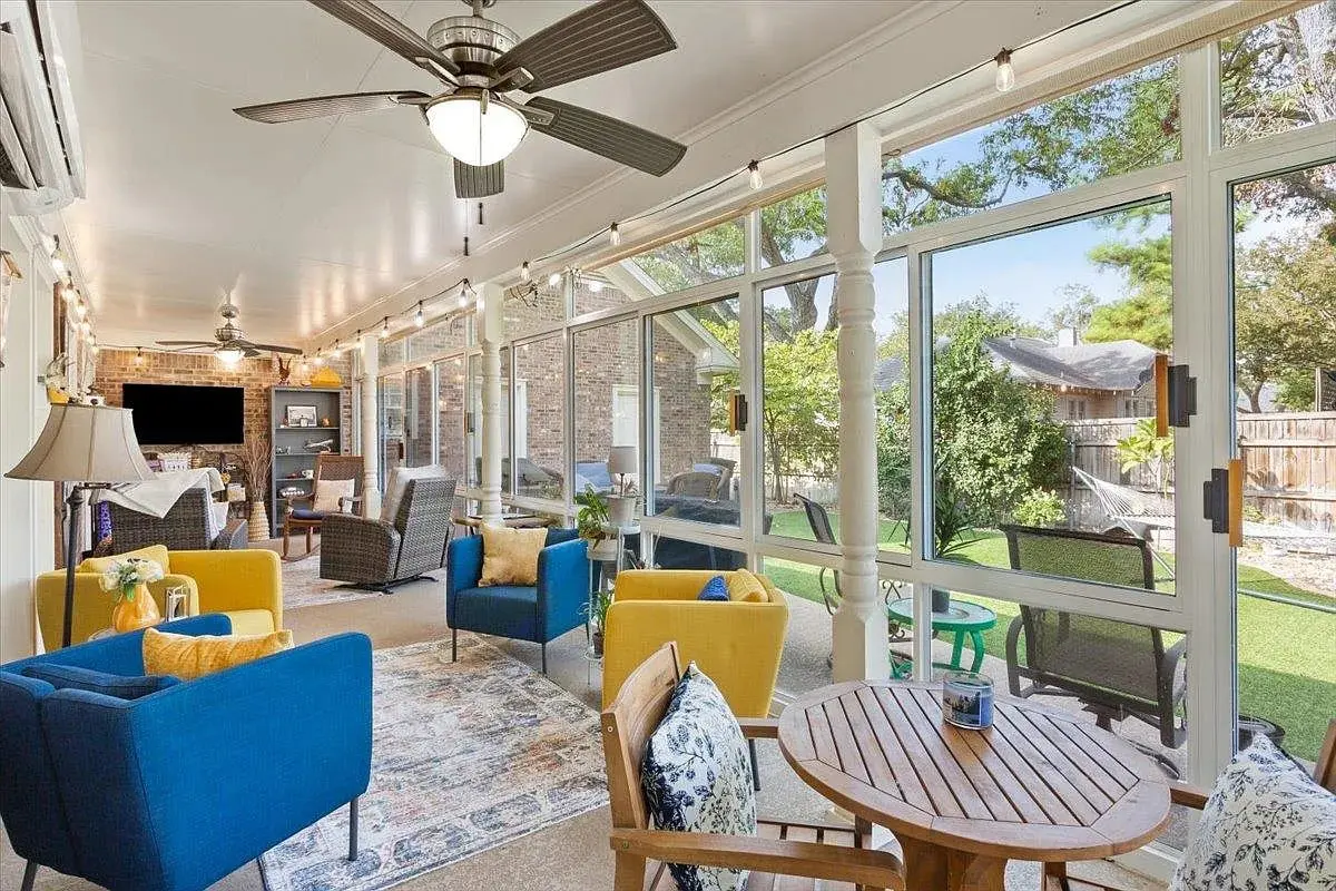 Sunroom with blue and yellow chairs, wooden table, ceiling fans, and large windows overlooking a garden.