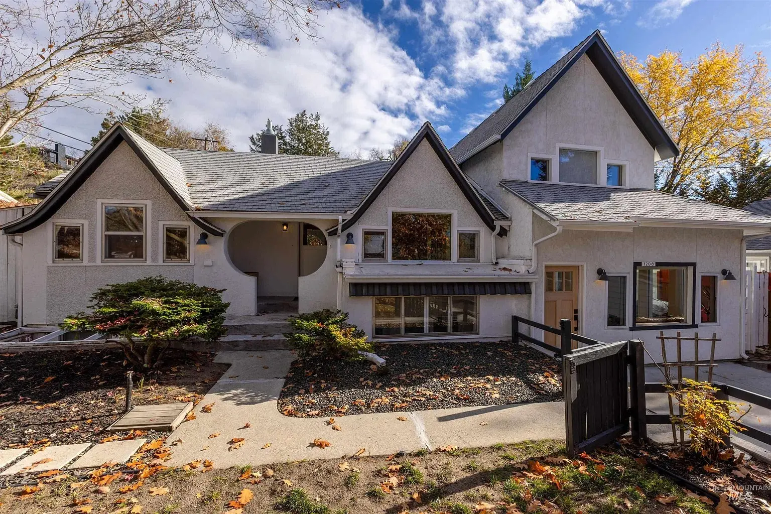 A two-story house with a unique roof design, surrounded by autumn trees and a small garden.