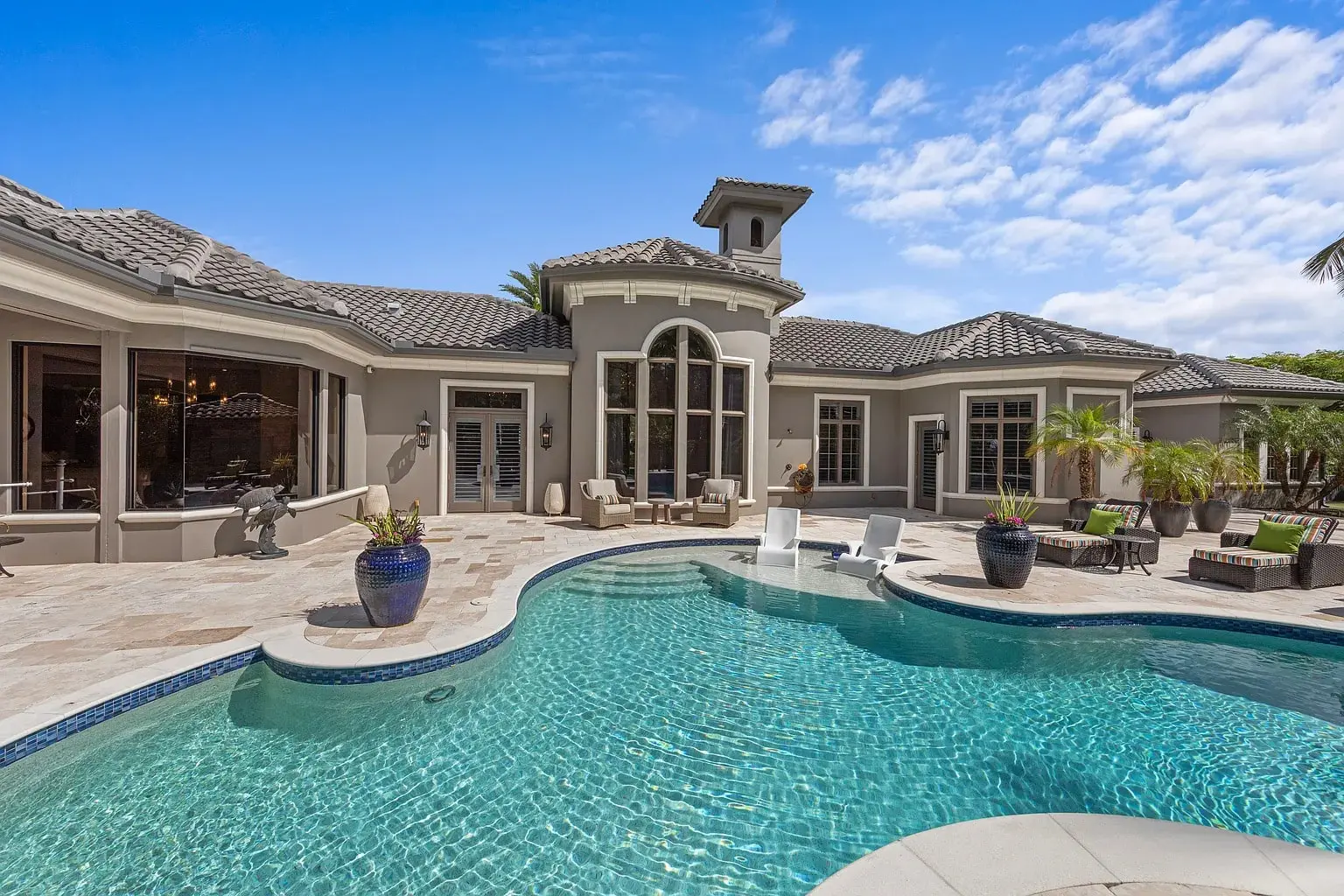 Luxurious house with a swimming pool, patio furniture, and palm trees under a blue sky.