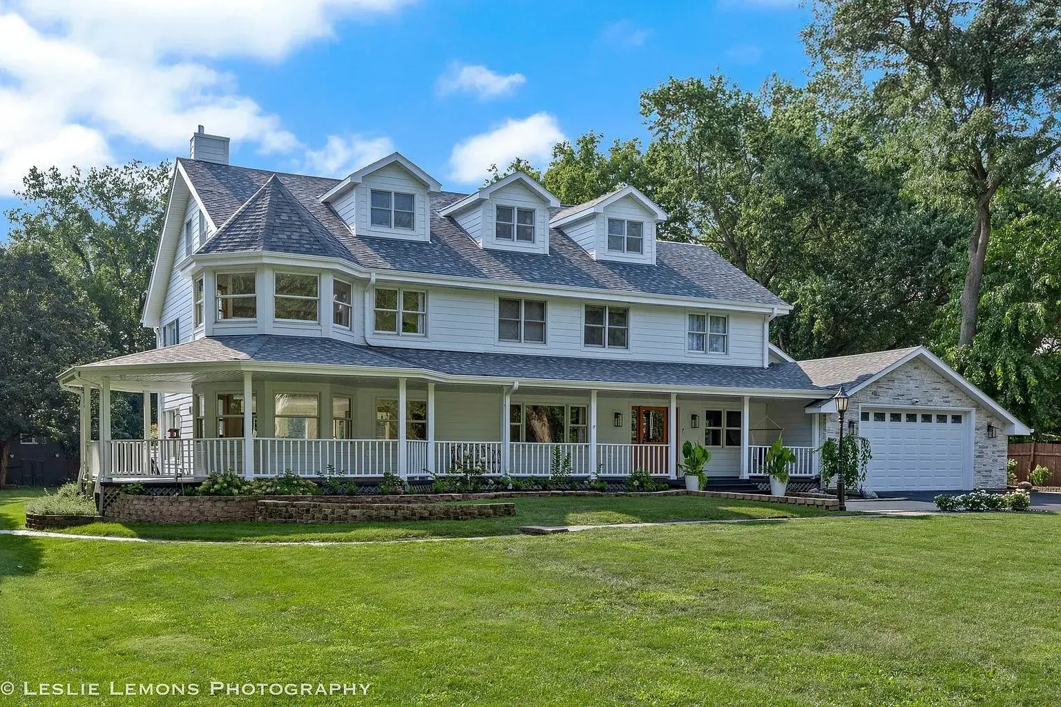 Large white two-story house with a wraparound porch and attached garage, surrounded by greenery.