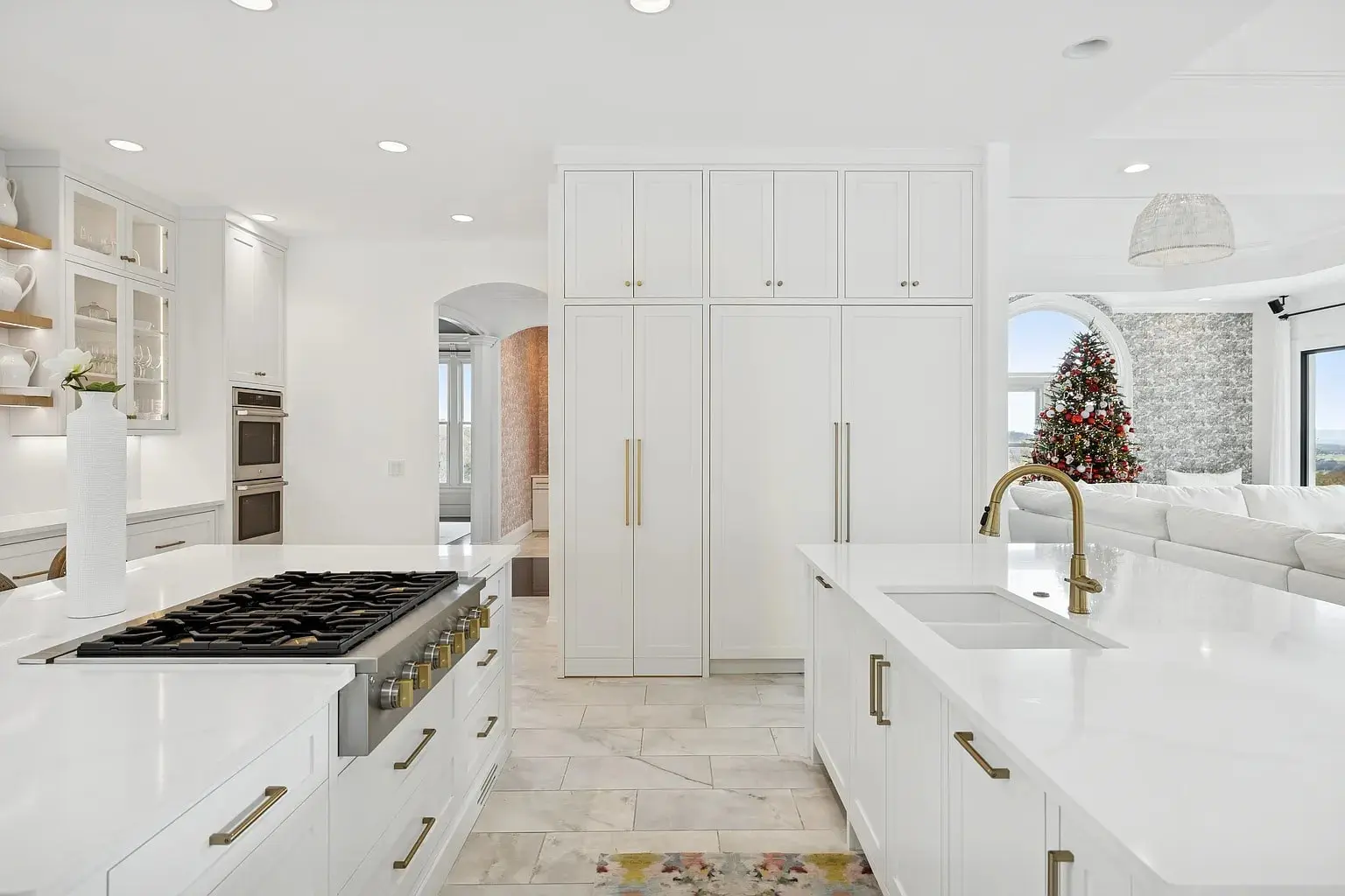 Modern white kitchen with island, gold fixtures, and a view of a living room with a Christmas tree.