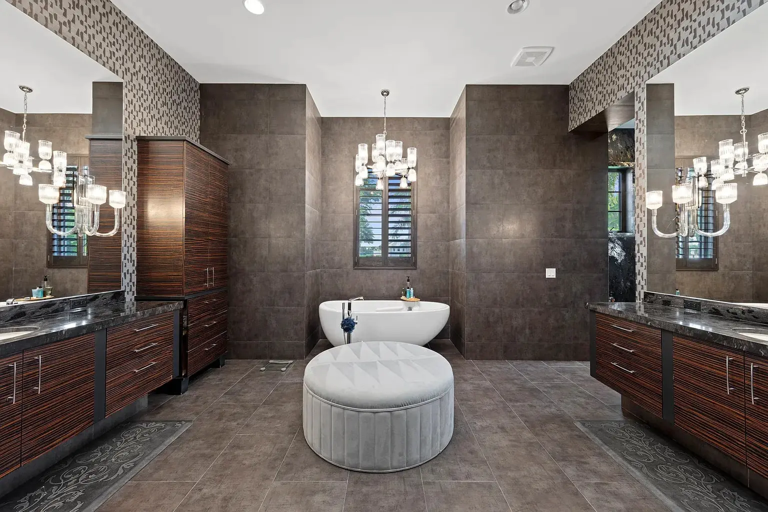 Luxurious bathroom with a central bathtub, chandeliers, dark wood cabinets, and a round ottoman.