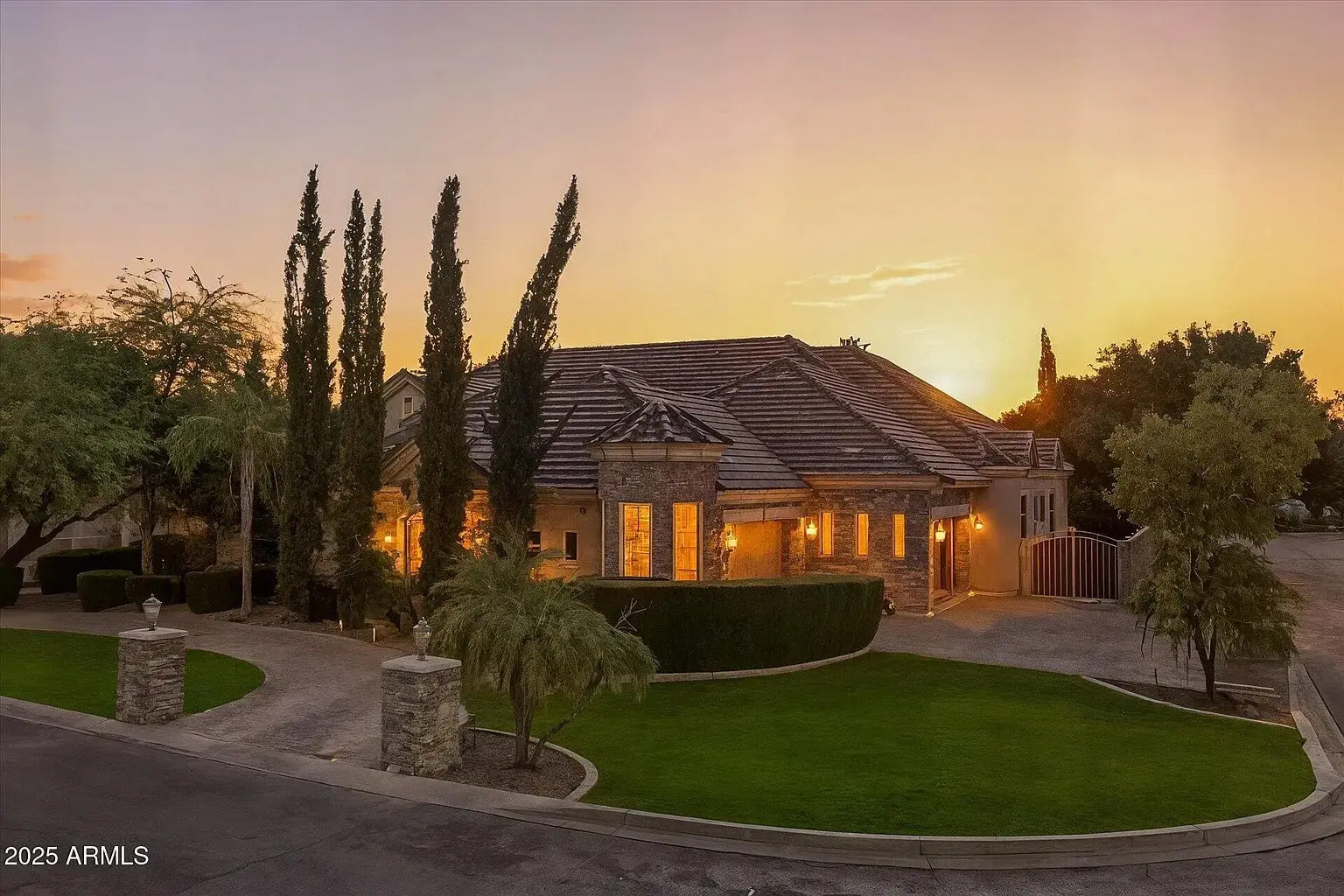 Large stone house with a tiled roof, surrounded by trees and a well-maintained lawn at sunset.