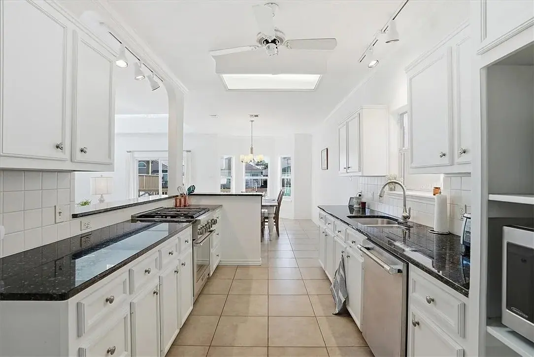 Spacious white kitchen with black countertops, tile floor, and ceiling fan, leading to dining area.