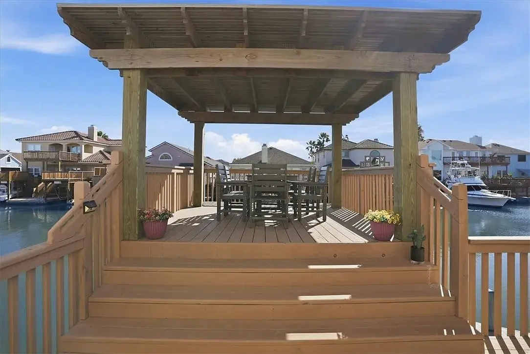 Wooden gazebo with table and chairs overlooking a waterfront with houses and boats.