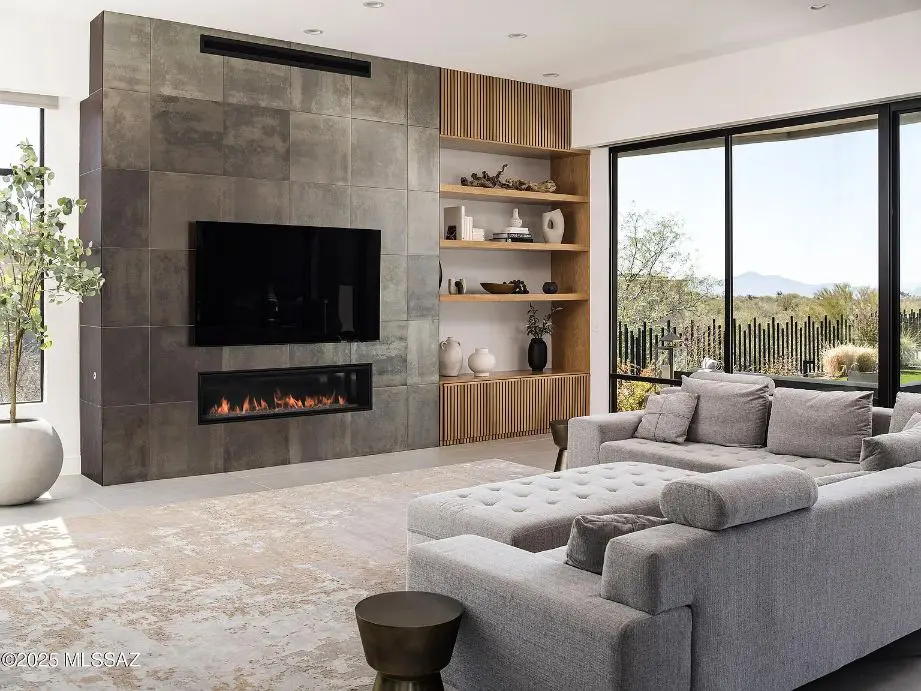 Modern living room with a gray sectional sofa, wall-mounted TV, fireplace, and large windows with a view.