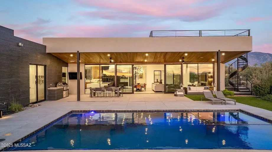 Modern house with large glass windows, pool, patio seating, and mountain views at sunset.