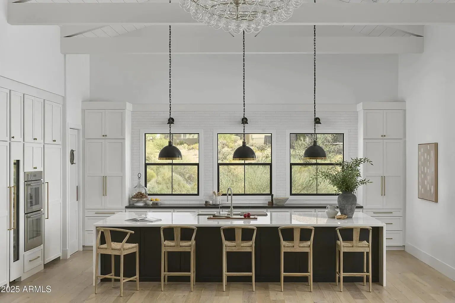 Modern kitchen with a large island, wooden stools, pendant lights, and three windows with outdoor view.
