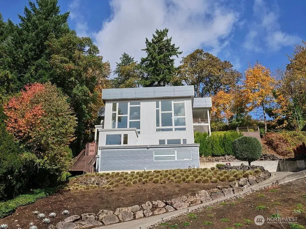Modern two-story house with large windows, surrounded by trees and a landscaped yard.